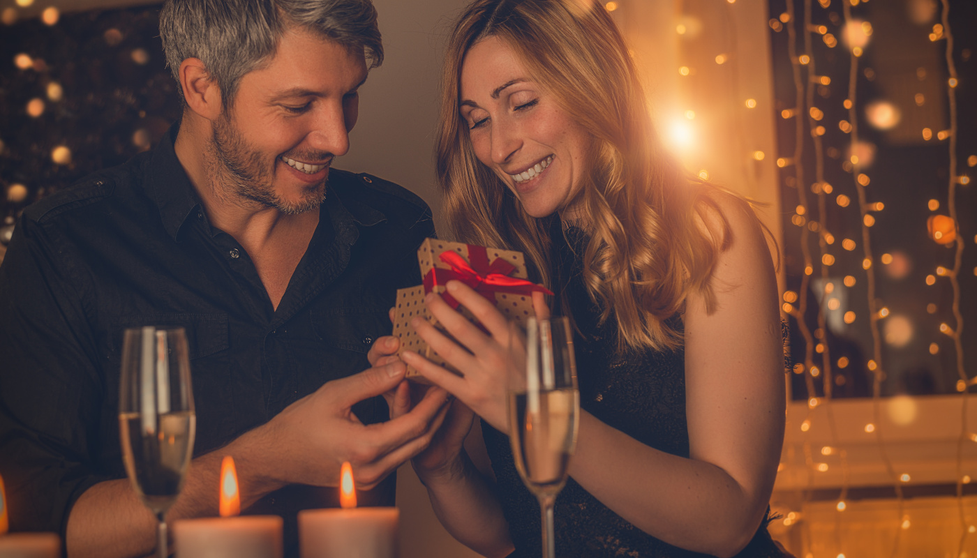 Ein junges Paar sitzt lächelnd bei Sekt und Kerzenlicht zusammen. Sie öffnet ein Geschenk.
